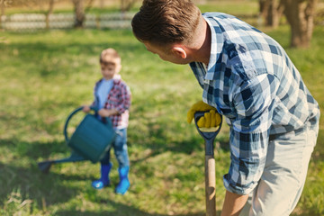 Love for work. Close up of an adult man digging the ground while his little son holding a watering can