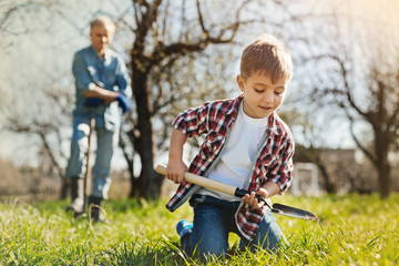 Enjoy doign it. Little positive boy sitting on the grass and digging the ground with a spade while his father standing in the background