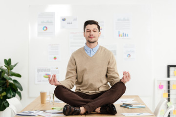 relaxed businessman with eyes closed sitting in lotus position on table in office