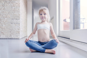 Little blond girl doing a yoga exercise in the room near the window