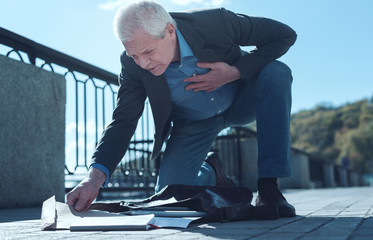 Need to take care of myself. Low angle shot of a senior man suffering from a violent paint in a heart area bending down and picking his stuff after dropping his bag in a pain.