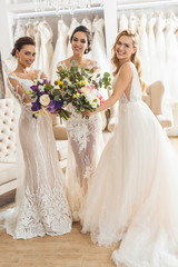 Naklejka premium Happy women in wedding dresses with flowers in wedding salon