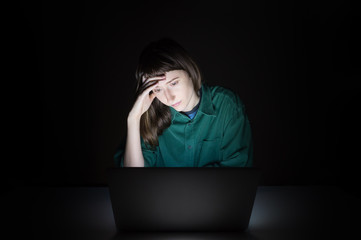 Tired and exhausted young woman at laptop computer indoors late in the evening. Portrait of thoughtful female student or worker sitting in front of computer screen at night, concept of anxiety