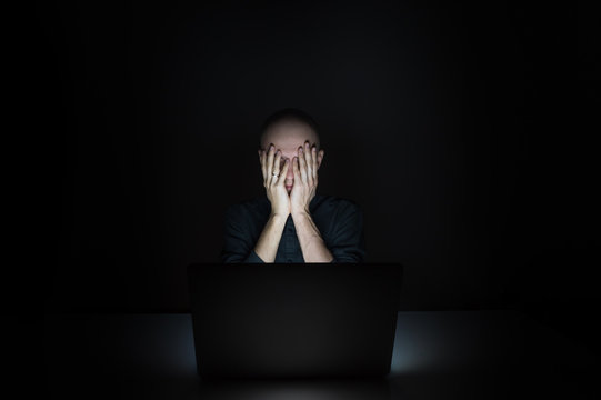 Tired And Exhausted Young Man At Laptop Pc Late In The Evening. Portrait Of Depressed Male Student Or Worker Sitting In Front Of Computer Screen At Night, Hiding Face In Hands - Concept Of Overworking