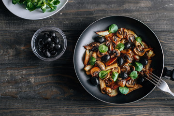 Fried potatoes with mushrooms, olives and greens in a frying pan on a wooden table vegetarian food top view
