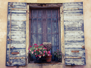Fototapeta na wymiar Colorful daisy flowers and cactus in pots placed on the window of the old stone house. Closed window with lace curtain, rusty lattice and open wooden shutters. (Arles, Provence, France) Toned image.