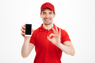 Excited happy young delivery man showing display of mobile phone make okay gesture.