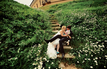 Bride and groom sitting on steps in chamomile flowers old yellow castle background at wedding Day summer nature. Bridal couple, Happy Newlywed woman and man embracing in green park. Honeymoon