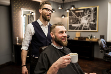 Happy barber and customer drinking coffee in salon
