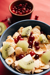 Delicious crispy cornflakes with kiwi pieces, almonds and cranberries in bowl, healthy breakfast on red background