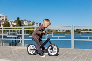 Happy four years old boy riding bicycle without pedals on the background of river and buildings. Sport concept. First balance bike for little children. Active and fun childhood outdoors