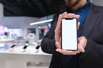 A man in a suit holds in his hands a modern smartphone with a white screen. A business man shows a trendy smartphone against the backdrop of an electronics store. The consultant offers a smartphone