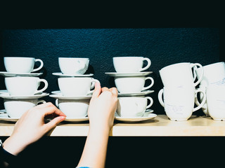 group of white coffee cup arrange on wooden shelf in coffee shop by beauty woman hand with black wallpaper background