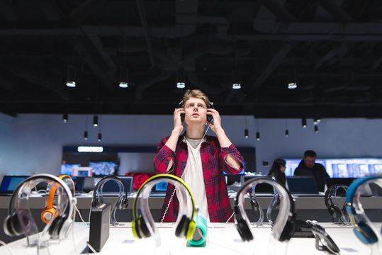 A Young Man Chooses A Headset In A Modern Store. Headphone Test. Portrait Of A Man In The Store While Listening To Music In The Headphones