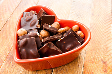 Red heart shaped bowl with chocolate pieces and nuts is standing diagonally on wooden table.