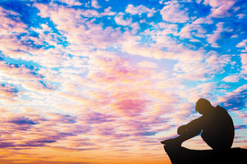 Silhouetted photo. Lonely man  sitting on cliff. He is unhappy and sad. He is watching the evening...