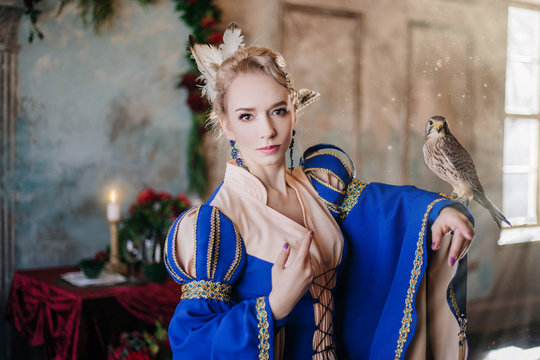 Beautiful girl in a medieval dress in blue. A model with white hair, a hairstyle with feathers. Bird of the falcon on the hand. Fantasy Photo