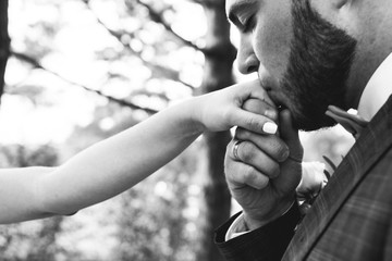 Stylish bearded groom in checkered suit is kissing bride's hand. Wedding couple in the forest. Romantic rustic photo outdoors. Beautiful love story details.