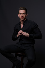 a guy in a black shirt and black pants sits on a dark chair, on a gray background