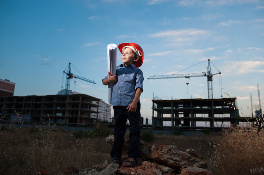 cute little boy in helmet with paper of draft in his hand on construction site with crane and new buildings