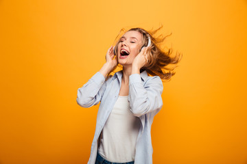 Photo of satisfied smiling woman 20s wearing braces in basic clothing expressing delight while listening to music on wireless earphones, isolated over yellow background