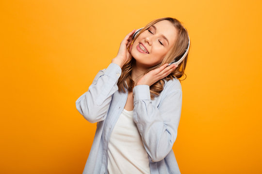 Picture of cute caucasian woman 20s wearing braces in casual clothing listening to music using wireless earphones, isolated over yellow background