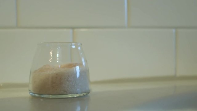 fingers pinch some bath salt from a clear glass container on the side of a tub and drop the salt in the bathtub.
