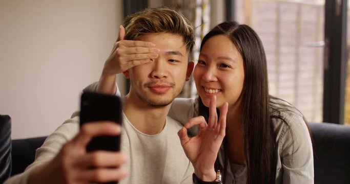 4K Cute Asian couple posing for funny selfies with smartphone at home