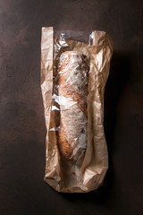 Loaf of fresh baked artisan whole grain ciabatta bread in market paper bag over dark brown texture background. Top view, copy space.