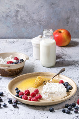 Ceramic plate of homemade cottage cheese served with blueberries, raspberries, bottle of milk and honeycombs over white marble texture table as background.