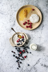 Ceramic bowl of homemade cottage cheese served with blueberries, raspberries, bottle of milk and honeycombs over white marble texture background. Top view, space