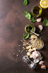 Traditional Basil pesto sauce in glass jar with ingredients above fresh basil, olive oil, parmesan cheese, garlic, pine nuts, lemon over dark texture background. Top view, space
