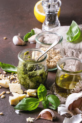 Traditional Basil pesto sauce in glass jar with ingredients above fresh basil, olive oil, parmesan cheese, garlic, pine nuts, lemon on cloth over dark texture background. Close up