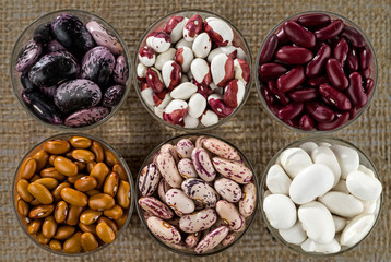 Six different varieties of beans in round glassware on the background of the texture of coarse cloth.