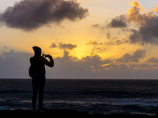 Girl standing at the Atlantic coast captering the sunset with a smartphone.