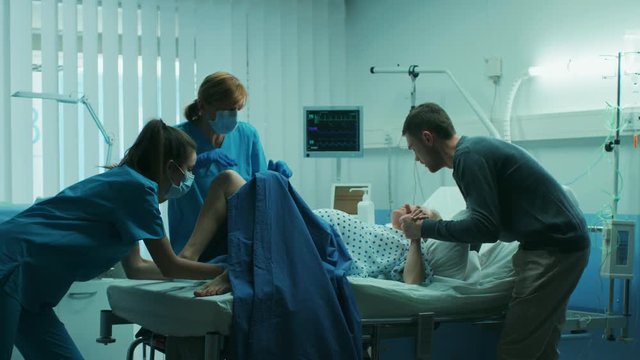 In the Hospital Woman in Labor Pushes to Give Birth, Obstetricians Assisting, Spouse Holds Her Hand. Modern Maternity Hospital with Professional Midwives. Shot on RED EPIC-W 8K Helium Cinema Camera.