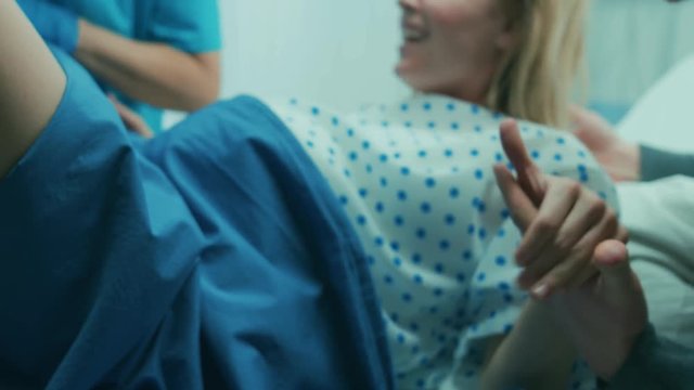 In the Hospital Woman in Labor Pushes to Give Birth, Tense Moment She Grabs Sheets in Pain. Face Close-up, Obstetricians Assist, Husband Supports. Shot on RED EPIC-W 8K Helium Cinema Camera.
