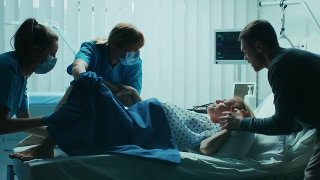 In the Hospital Woman in Labor Pushes to Give Birth, Obstetricians Assisting, Husband Holds Her Hand. Modern Delivery Ward with Professional Midwives.  Shot on RED EPIC-W 8K Helium Cinema Camera.