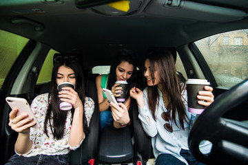 Beautiful three women is sitting and driving a car holding a mobile phone and messaging. Three girls drinking coffee and looking forward happily.