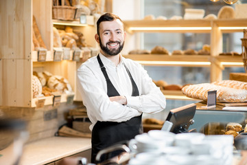 Portrait of a handsome seller in uniform standing in the small and beautiful store with bakery...
