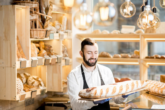 Handsome seller in uniform holding a big loaf of bread in the store with bakery products