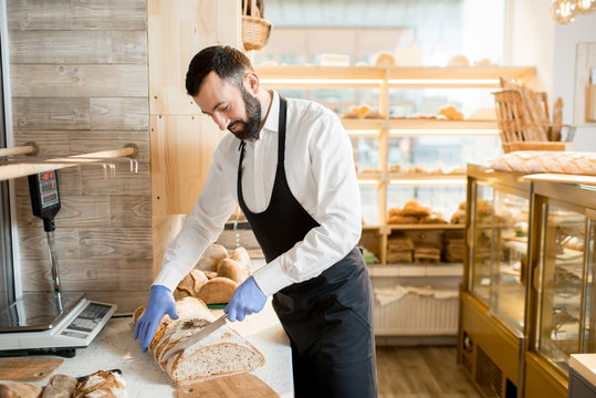 Man seller cutting fresh bread for selling in the store with bakery products