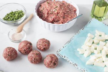 Marble table top with ingredients to make meatballs including raw ground beef, mozzarella cheese, chives and salt