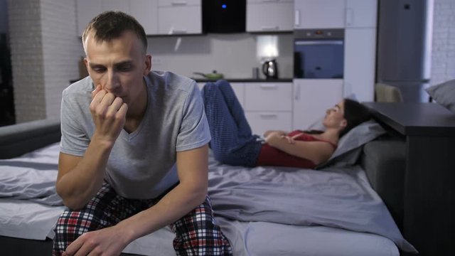 Young mid adult couple having problems in relationship. Woman lying in bed while her pensive frustrated man sitting facing the camera. Married couple thinking about their argument and sexual problems