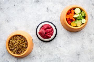 pet care with dry food, vegetables and raw meat for dog or cat in plastic bowl on stone background top view space for text