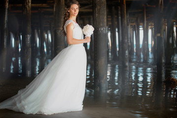 Gorgeous artistic portrait of beautiful bride in white dress holding bridal bouquet under pier