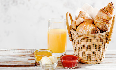 Baking breakfast croissants and bun in basket with orange juice, honey, butter, raspberry jam on white wood table