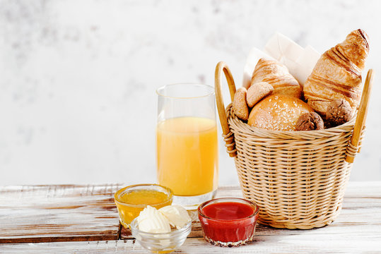 Baking breakfast croissants and bun in basket with orange juice, honey, butter, raspberry jam on white wood table