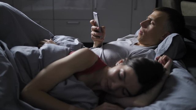 Young mid adult couple lying in bed at night under blanket. Young wife sleeping, while her handsome husband texting and cheating on her online via mobile phone.