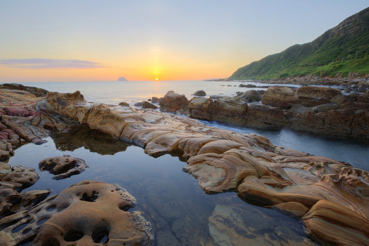 Beautiful scenery of dawning sky by rocky beach in northern Taiwan with reflection of golden sun light on tranquil sea water ~ Romantic view of sunrise by sea shore (long exposure effect) 